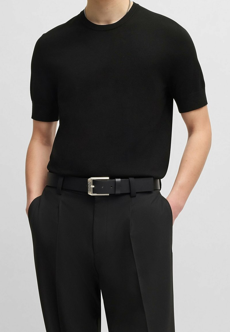 Pull à manches courtes noir à la texture lisse, associé à un pantalon noir sur mesure et une ceinture noire avec une boucle argentée.