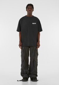 Tricou oversized negru cu un logo alb pe piept, asortat cu pantaloni cargo negri largi, având multiple buzunare și tivuri cu siret.