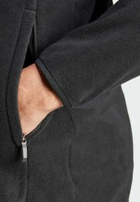 Black fleece jacket with a textured surface, featuring a zippered pocket and contrasting trim along the cuffs.