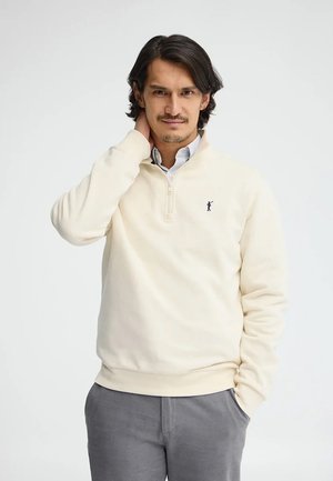 Mężczyzna o ciemnych włosach, ubrany w kremowy sweter z półzamkiem i szare spodnie, stoi z jedną ręką w kieszeni, a drugą dotyka szyi.