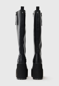 Fekete, térd fölé �érő csizmák sima bőrfelülettel, bordázott díszítéssel a bokánál, vastag platformtalpakkal és oldalsó cipzárokkal a könnyű felvétel érdekében.