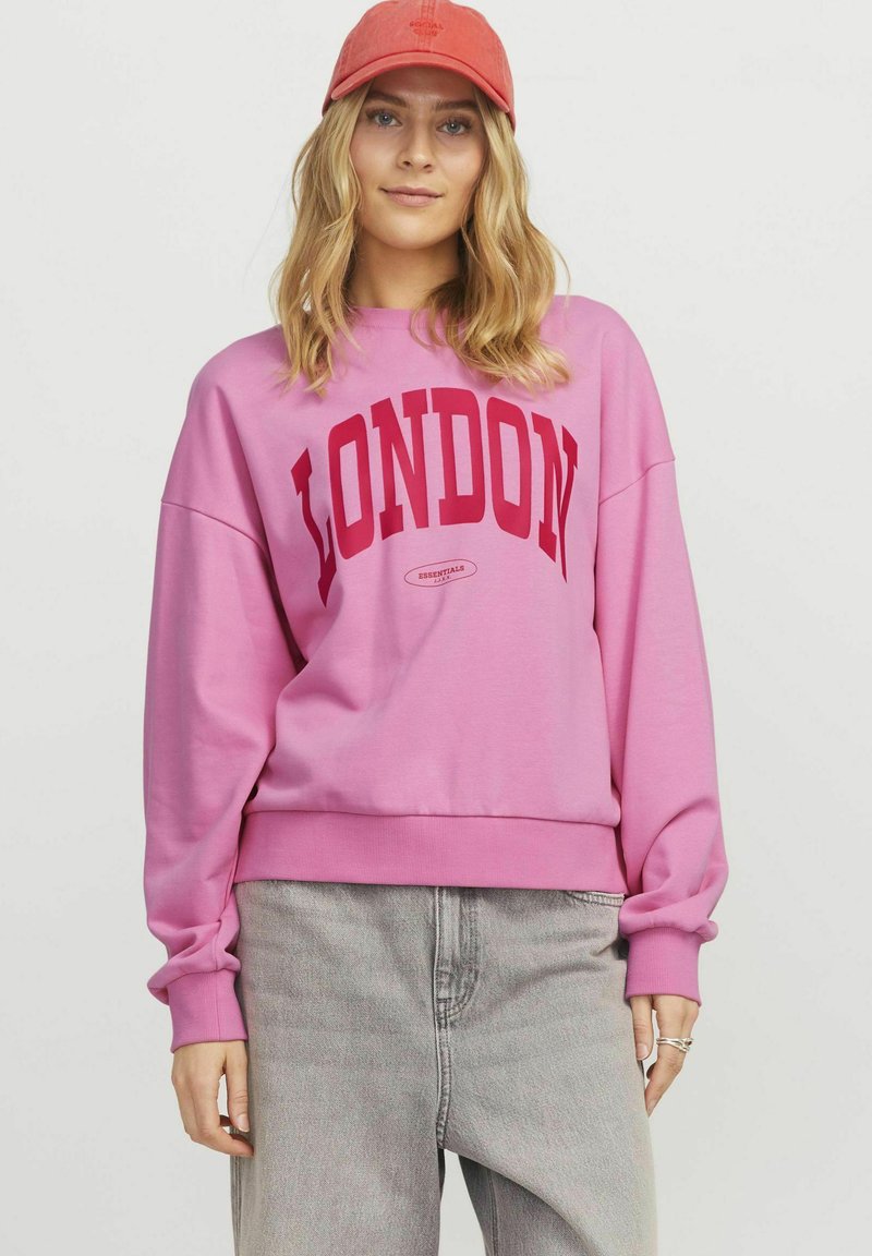 Sweatshirt rose avec "LONDON" en lettres rouges audacieuses, à manches longues et col rond, associé à un jeans gris et une casquette rouge.