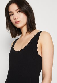 Sort ribbet top med bølget udskæring og stropper. Har en t�ætsiddende silhuet og tekstureret stof for ekstra detalje.