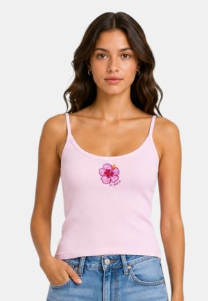 Donna con lunghi capelli ondulati che indossa un top rosa chiaro con spalline sottili, con un fiore rosso e la scritta "St. Barth", abbinato a jeans blu.