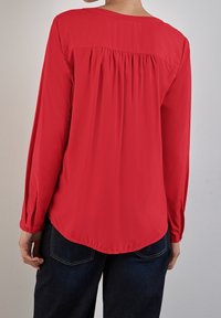 Rote Bluse mit langen Ärmeln, abgerundetem Saum, Falten an der Rückseite, weicher Textur und geknöpften Manschetten. Kombiniert mit dunklen Jeans.