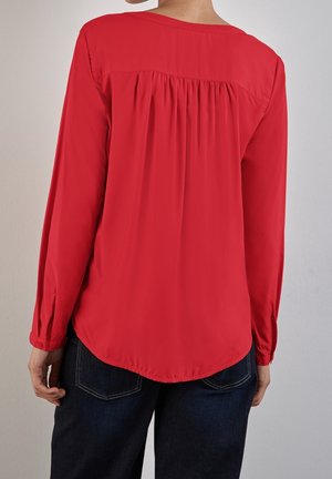 Blusa rossa a maniche lunghe con orlo arrotondato, pieghe sul retro, texture morbida e polsini con bottoni. Abbinata a pantaloni in denim scuro.