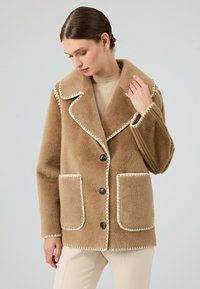 Tan wollen jas met een brede kraag, contrasterende crèmekleurige stiksels, twee zakken aan de voorkant en zwarte knopen. Zacht, gestructureerd materiaal met een relaxte pasvorm.