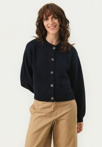 Navy blue knitted cardigan with a round neckline, seven buttons, and two front pockets; features ribbed cuffs and hem.