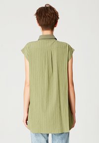 Tunique à manches courtes olive verte avec un tissu texturé, un col boutonné et un pli à l'arrière, associée à un jean en denim bleu clair.