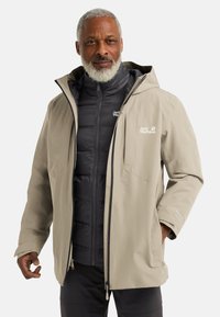 Veste imperméable beige avec une capuche, présentant une couche intérieure isolante noire. Plusieurs poches zippées et logos sur les deux couches.
