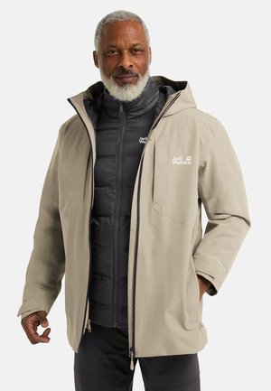Veste imperméable beige avec une capuche, présentant une couche intérieure isolante noire. Plusieurs poches zippées et logos sur les deux couches.