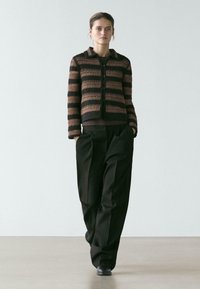 Frau trägt eine braun-schwarz gestreifte Strickjacke, schwarze weite Hosen und schwarze Schuhe, geht mit den Händen in den Taschen.