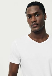 Fehér pamut póló V-nyakkal, rövid ujjakkal és sima textúrával. Közelkép a felső testrészről, amely bemutatja a nyakkivágást és az illeszkedést.