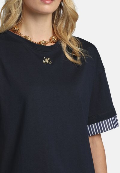 Blonde woman wearing a black T-shirt with a striped sleeve cuff and a gold chain necklace with large knots.