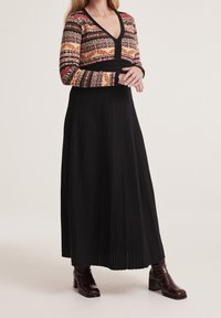 Dlouhá černá plisovaná šaty s barevným vzorovaným topem s dlouhým rukávem a výstřihem do V, který má těsně přiléhající korzet. Hnědé boty na podpatku.