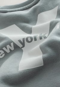 Materialul bluzei gri este moale, cu o textură fină, având un logo alb mare și textul "new york" în gri închis. Are guler rotund și un design casual.