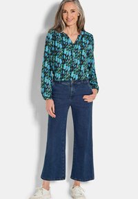 Blouse fluide noire avec un motif abstrait vert et bleu, assortie à un jean en denim foncé à jambes larges et des baskets blanches.