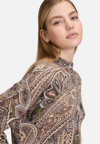 Młoda kobieta z prostymi blond włosami, ubrana w bluzkę z wysokim kołnierzem, zdobioną szczegółowymi brązowymi, beżowymi i szarymi wzorami paisley na białym tle.
