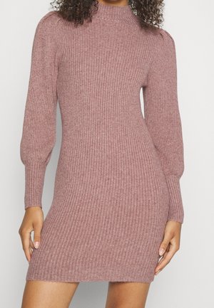 Robe en maille côtelée rose poudrée ajustée avec de longues manches bouffantes et un col montant, portée par une personne debout aux cheveux bouclés, les mains reposant sur les côtés.