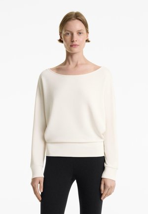 White, long-sleeve top in a soft, lightweight fabric with a wide neckline and fitted hem, paired with black leggings.