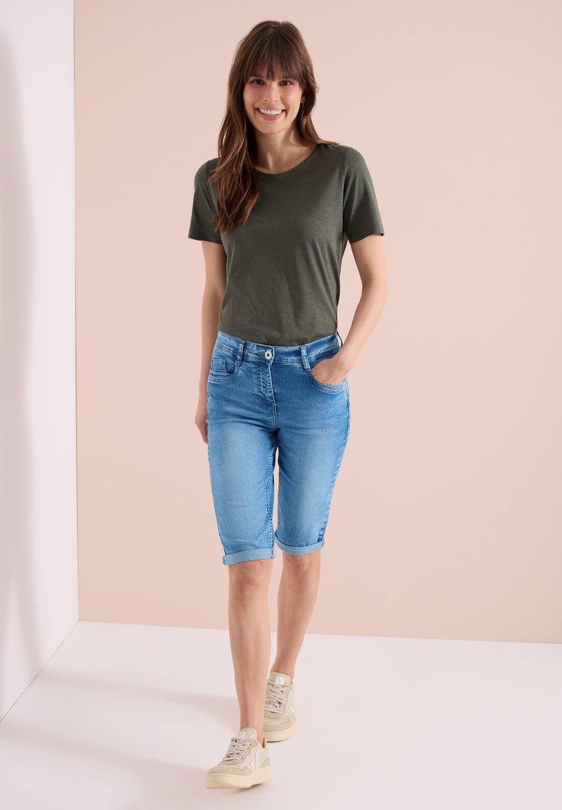 Femme souriante, portant un t-shirt vert olive, un short en jean bleu retroussé et des baskets beiges, debout devant un fond beige.