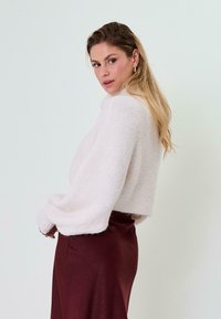 Trumpa, šviesiai rožinė, pūkuota megztinė su plačiomis rankovėmis ir aukšta apykakle, derinama su bordo spalvos sijonu prieš šviesų foną.