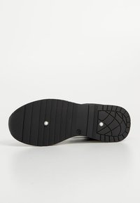 Semelle de chaussure noire avec des motifs de bande de roulement horizontaux et géométriques et deux petits rivets métalliques visibles sur fond blanc.