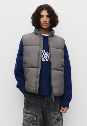Grijze gewatteerde vest met een hoge kraag, gedragen over een blauwe gebreide trui. Voorzien van ritssluiting en een gladde textuur. Casual, lichtgewicht ontwerp.