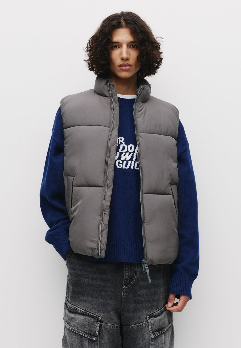 Graue gepolsterte Weste mit hohem Kragen, getragen über einem blauen Strickpullover. Verfügt über einen Reißverschluss und eine glatte Textur. Lässiges, leichtes Design.