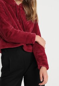 Hoodie cropped in finto pelliccia rossa con coulisse, caratterizzato da maniche lunghe e una texture morbida, abbinato a pantaloni neri.