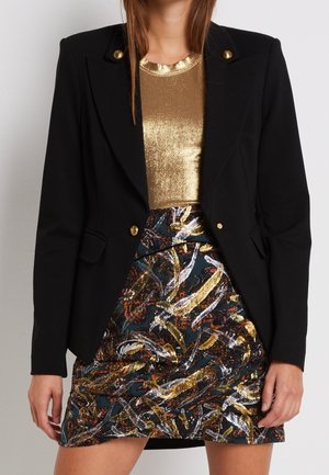 Nainen musta bleiseri, jonka napit ovat kultaiset, metallinhohtoinen kultaiseen vivahtava paita ja monivärinen kuviollinen hame, jossa on kultaisia, hopeisia ja ruskeita yksityiskohtia.