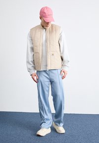 Colete acolchoado bege, sweatshirt cinza, calças de treino azul claro e boné rosa. Detalhes notáveis: fecho de correr, patch do logótipo e tecido texturizado.