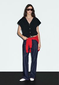 Schwarzes Kurzarmoberteil mit Knopfverschluss, roter Pullover um die Taille gebunden, dunkle Jeans mit weitem Bein, weiße Slipper und Sonnenbrille.