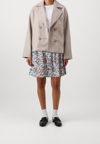 Donna che indossa una giacca beige doppiopetto, top bianco, gonna a fantasia floreale con fiori blu e arancio, calze bianche e mocassini neri.