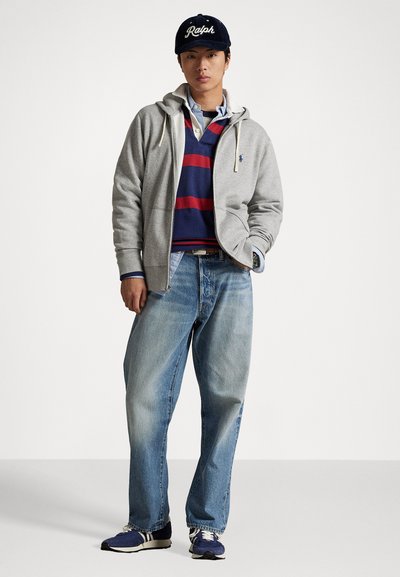 Jovem vestido com um boné azul marinho, um hoodie cinza com zíper, uma camisa listrada em azul e vermelho, jeans azul claro e ténis azul marinho, em pé com uma mão no bolso.