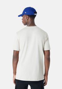 T-shirt en coton beige clair avec un col rond et des manches courtes, associé à une casquette bleu vif avec des accents de logo et des coutures contrastantes.