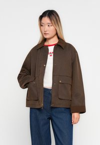 Jonge vrouw in een oversized bruine jas, witte crop top met rode letters en hooggesneden blauwe spijkerbroek, kijkend naar links tegen een effen achtergrond.
