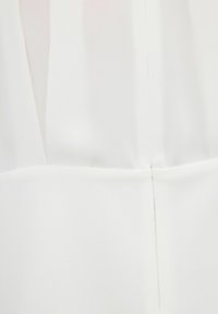 Détail de couture sur tissu blanc montrant les points de couture et un léger pli, mettant en valeur la construction du vêtement et la texture.