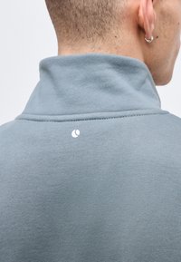 Στενό κοντινό πλάνο του λαιμού και αυτιού ενός ατόμου που φοράει ανοιχτό μπλε ρούχο με ψηλό γιακά και μικρό λευκό λογότυπο στο πίσω μέρος.