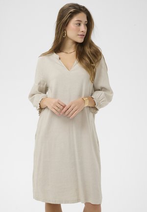 Žena s dlhými vlasmi, ktorá má na sebe voľné svetlo béžové šaty s tromi štvrťovými rukávmi, zlaté kruhové náušnice a zlatý náramok, pozerá sa bokom.