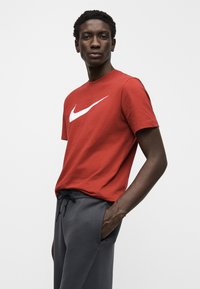 Tricou roșu din bumbac cu un logo mare alb Nike pe partea din față, asortat cu pantalonii sport gri cu șnur. Textură netedă, croială standard.