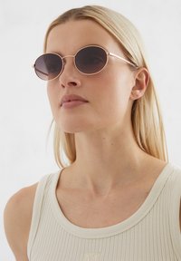 Lunettes de soleil rondes avec des verres teintés sombres, une fine monture métallique en or rose et un minimum de détails. Débardeur en tissu léger avec texture côtelée.