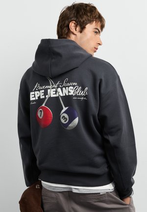 Jonge man met een zwarte hoodie met "EPE JEANS Club" en een afbeelding van biljartballen op zijn rug, houdt een bruine pet achter zijn rug.