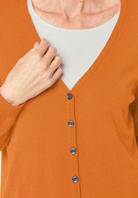Oranje V-hals cardigan gemaakt van gebreide stof, met marineblauwe knopen langs de voorkant en geribde manchetten. Close-up van een hand op de stof.