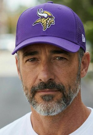 Mittelalterlicher Mann mit salz-und-pfefferfarbenem Bart, der eine lila Mütze mit einem Wikingerkopf-Logo und ein weißes Hemd trägt, draußen bei weichem Licht.