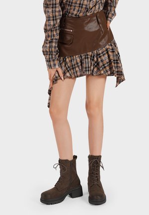 Donna che indossa una mini gonna in pelle marrone con orlo plaid arricciato, camicia a quadri coordinata e stivali alla caviglia marrone scuro con lacci, in piedi.