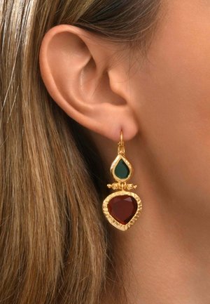 Boucle d'oreille en or avec une goutte verte et des pierres rouges en forme de cœur portée à l'oreille d'une personne aux cheveux châtain clair.