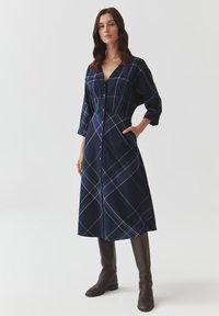 Robe à carreaux marine avec un devant boutonné, un décolleté en V et des manches trois-quarts, fabriquée en tissu doux avec des lignes vertes et violettes contrastantes.