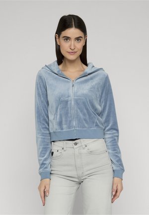 Donna con capelli scuri che indossa una felpa di velluto azzurro chiaro con zip e jeans a vita alta grigio chiaro, in piedi davanti a uno sfondo neutro.