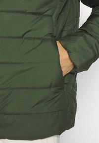 Giacca a vento verde con texture trapuntata, dotata di tasca laterale e design delle maniche aderente. Materiale leggero e resistente all'acqua.